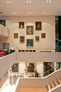 Photograph taken across an open-plan staircase to a balcony with a number of portraits of different sizes and two information panels.