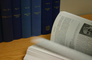 Image of a modern printed textbook on a desk in front of a set of bound reference works, with pages being turned.